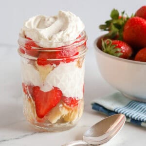 Shortcake parfait in a mason jar on marble countertop. A white bowl of strawberries in the background on top of a blue striped kitchen towel. A vintage teaspoon sits in front of the jar.