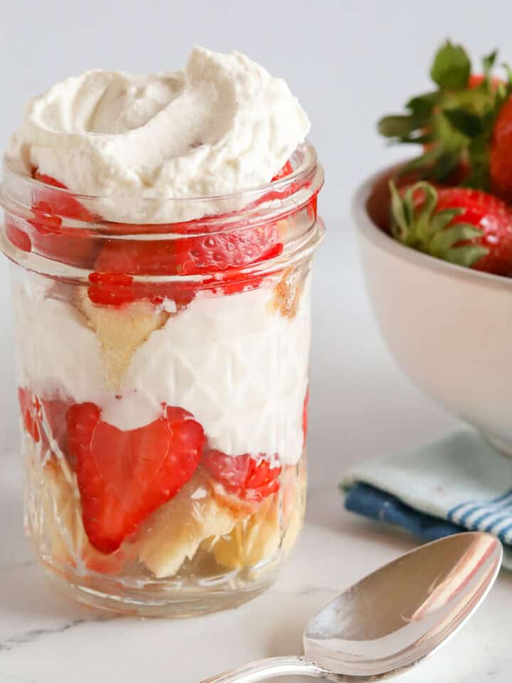 Shortcake parfait in a mason jar on marble countertop. A white bowl of strawberries in the background on top of a blue striped kitchen towel. A vintage teaspoon sits in front of the jar.