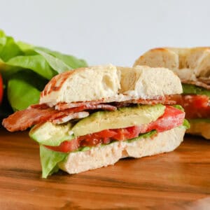 Half of bagel but on a wooden board with other half of sandwich and extra leaf lettuce in background.