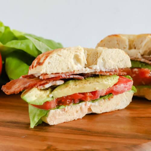 Half of bagel but on a wooden board with other half of sandwich and extra leaf lettuce in background.