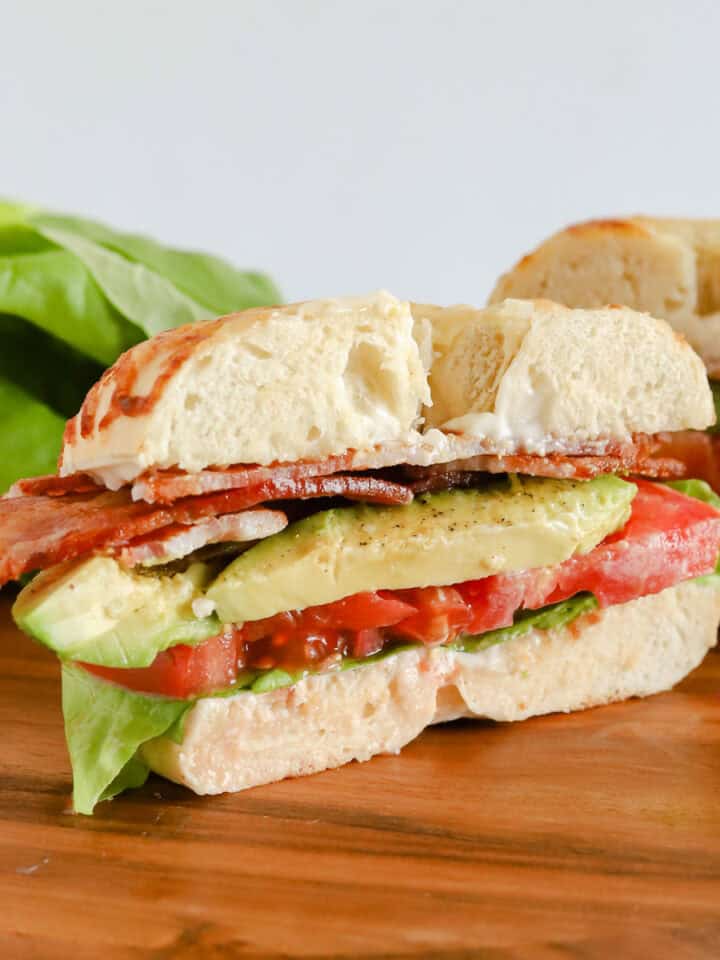 Half of bagel but on a wooden board with other half of sandwich and extra leaf lettuce in background.