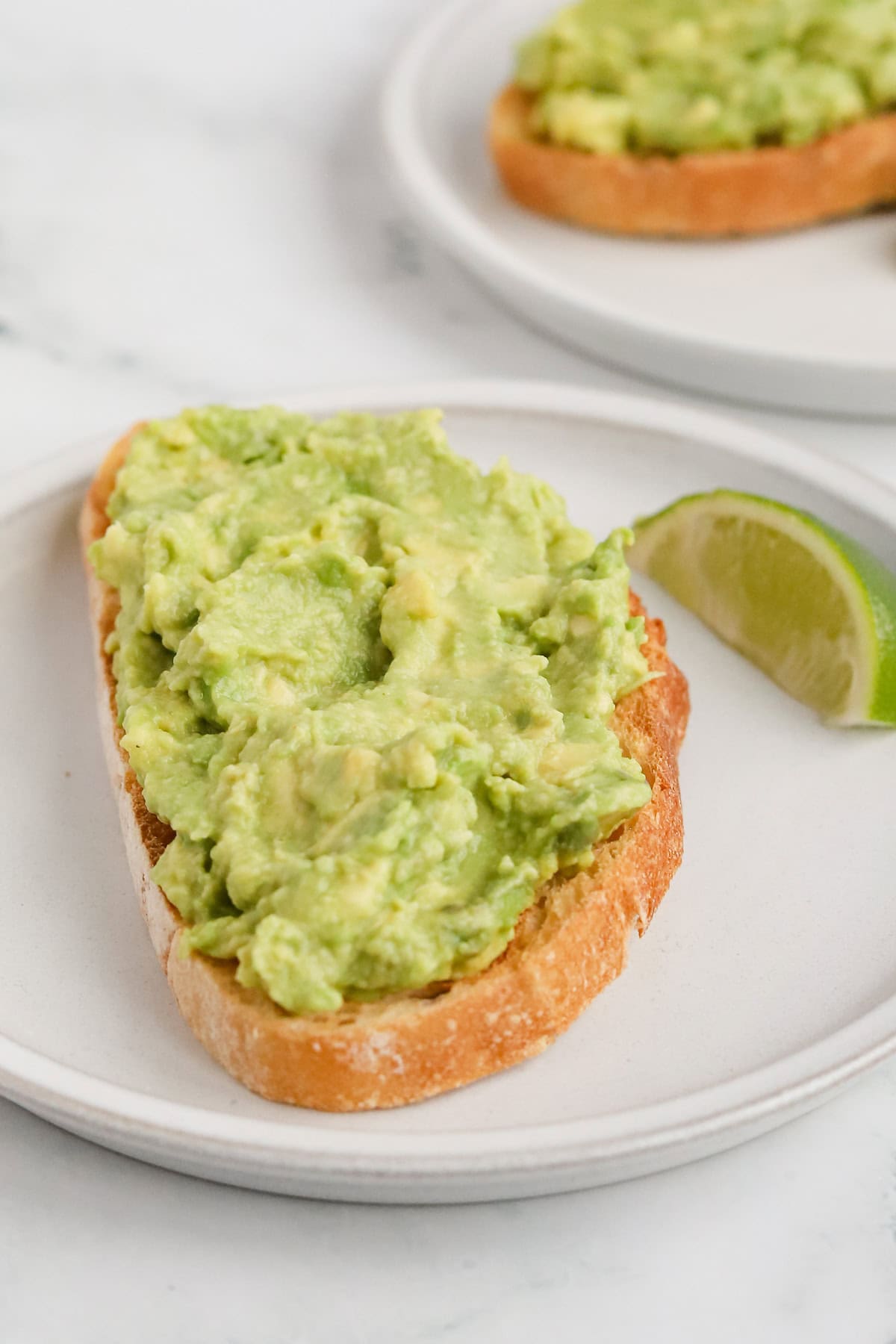 Piece of avocado toast on a white ceramic plate with a wedge of lime.