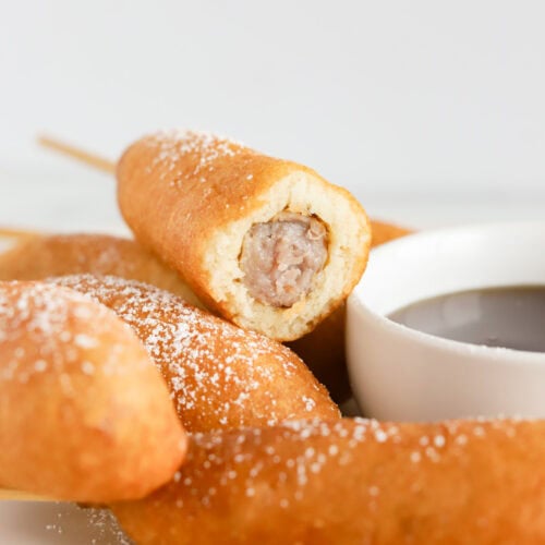 Corn dog with a bite taken out of end sitting on top of other corn dogs on a plate with a white bowl of maple syrup next to them.