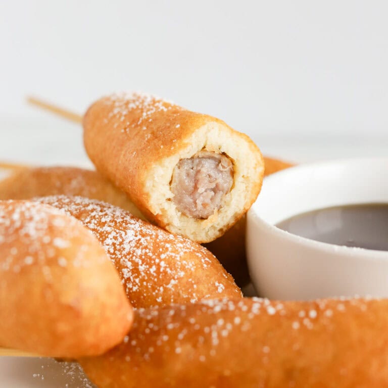 Corn dog with a bite taken out of end sitting on top of other corn dogs on a plate with a white bowl of maple syrup next to them.