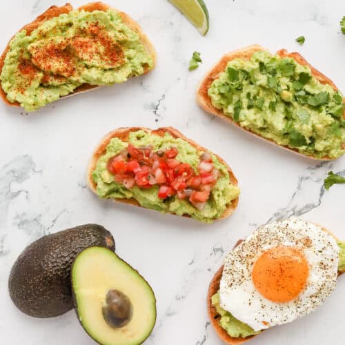 Four guacamole toast variations on marble countertop. A halved avocado also shown.