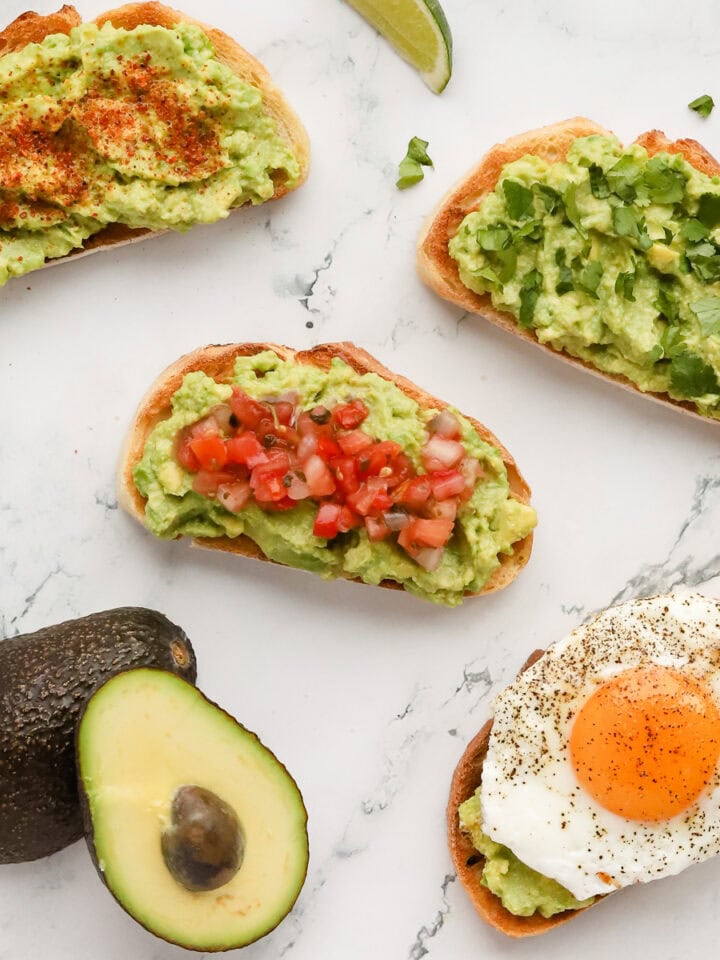 Four guacamole toast variations on marble countertop. A halved avocado also shown.