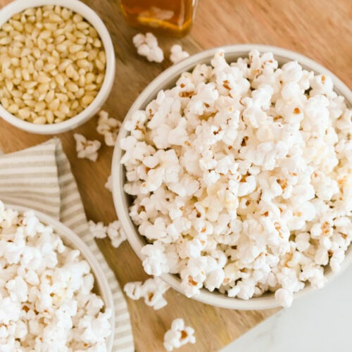 White bowls of truffle popcorn on a wooden cutting board. A smaller white bowl of kernels and bottle of truffle oil next to the bowl.