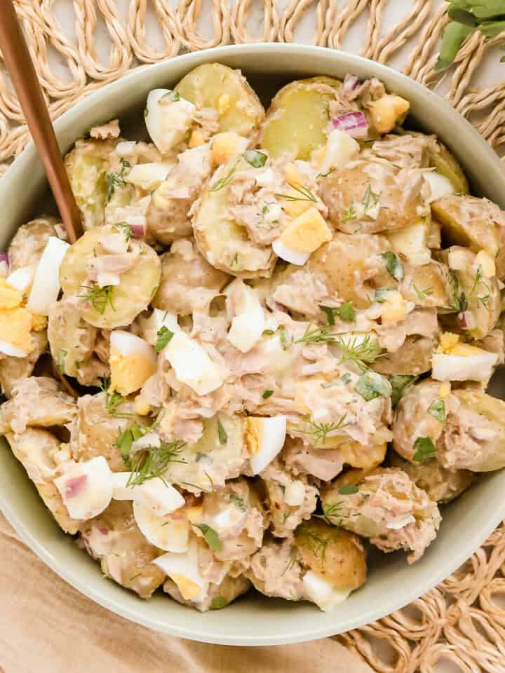 Tuna potato salad in a light green ceramic bowl on a wicker placemat with a jar of fresh herbs alongside.