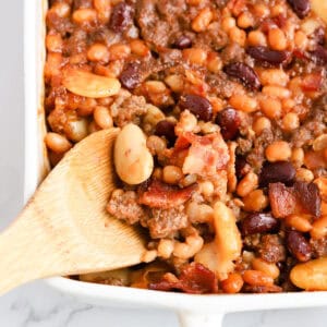 Wooden spoon scooping beans out of white baking dish.