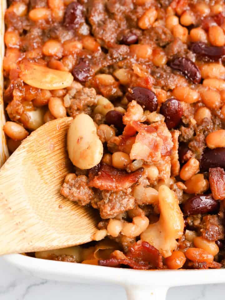 Wooden spoon scooping beans out of white baking dish.