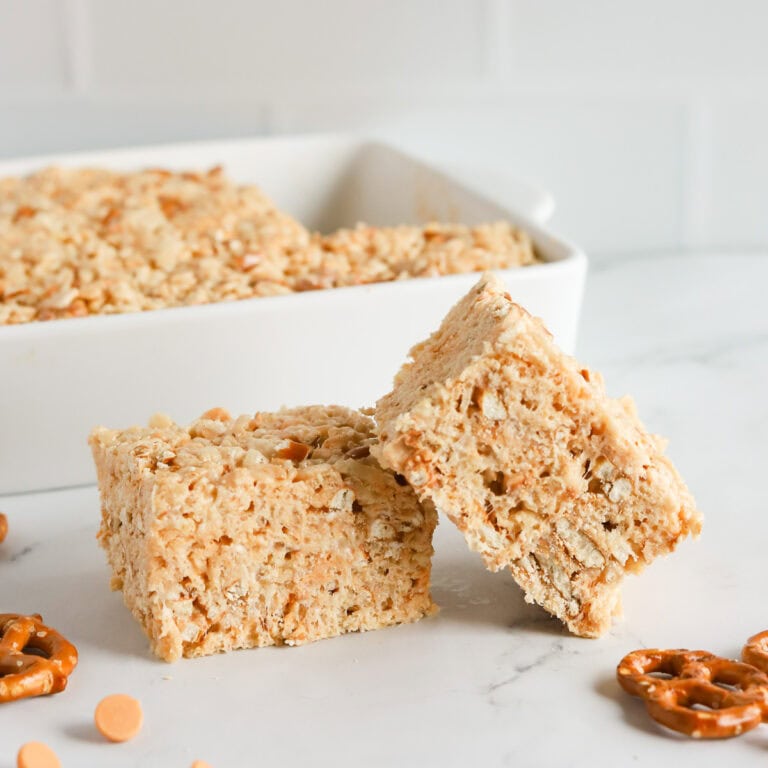 Two bars sitting on marble counter with pan of bars in the background. Whole pretzels and butterscotch chips scattered on counter.