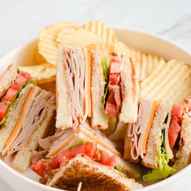 Clubhouse sandwich cut into triangles piled on a white plate with wavy potatoes chips in background.
