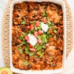 Walking taco casserole in white baking dish with shredded lettuce, diced tomato and 2 dollops of sour cream on top. Dish is sitting on wicker placemat with halved avocado in the corner.