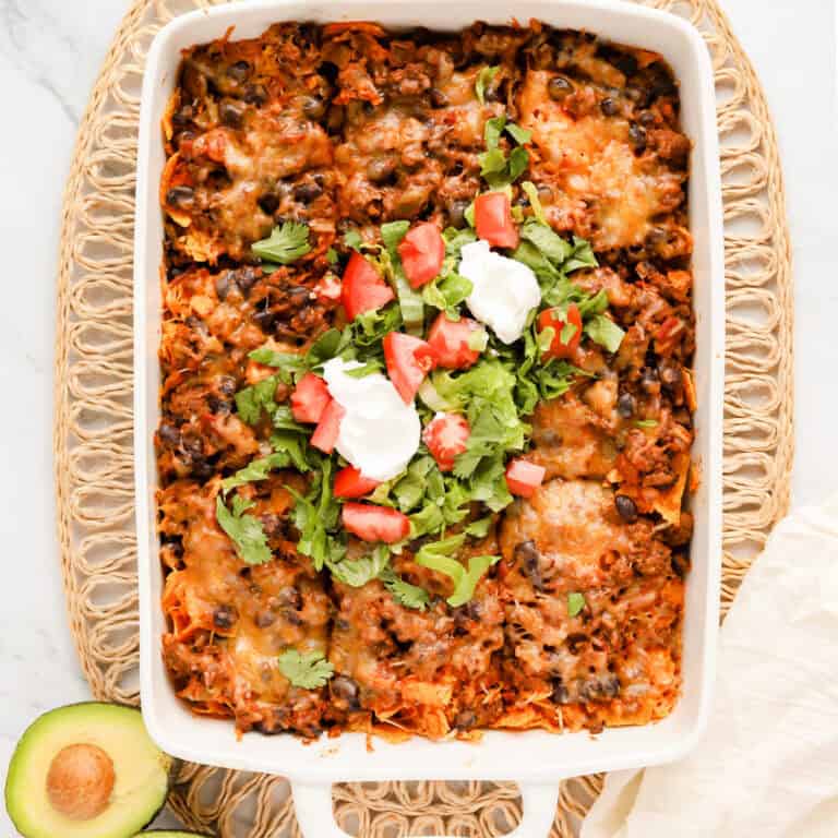 Walking taco casserole in white baking dish with shredded lettuce, diced tomato and 2 dollops of sour cream on top. Dish is sitting on wicker placemat with halved avocado in the corner.