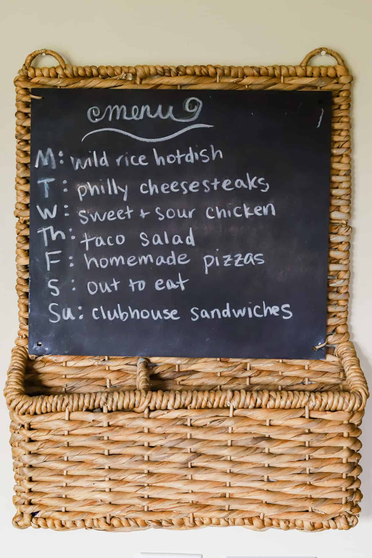 Wicker framed chalkboard hanging on wall with weekly dinner menu written on it.