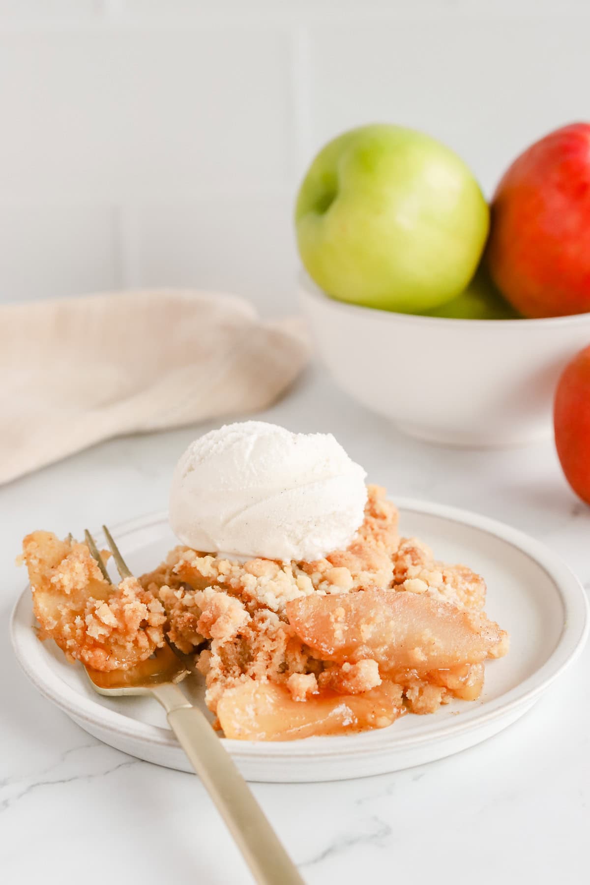 Serving of apple cobbler on a white ceramic plate with a scoop of vanilla ice cream on top. Bowl of apples are in the background.