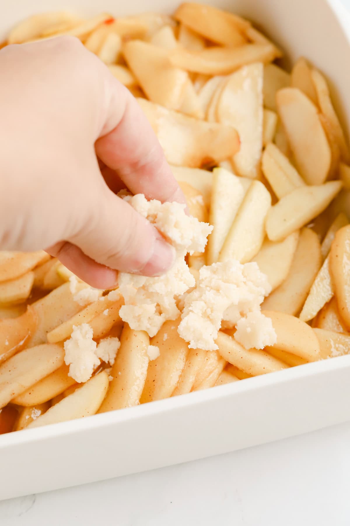 Hand placing cake mix cobbler topping on top of apples in a white baking dish.