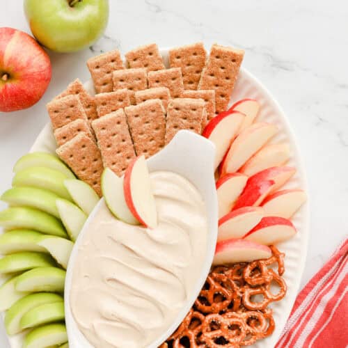 Square photo of apple dip on a platter with graham crackers, 2 kinds of apples and pretzel twists.