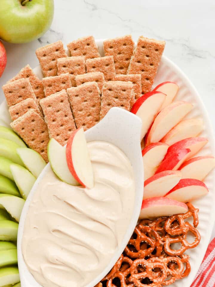 Square photo of apple dip on a platter with graham crackers, 2 kinds of apples and pretzel twists.