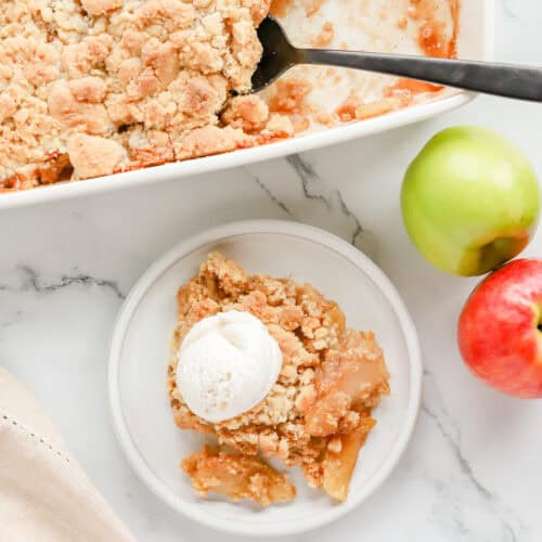Overhead shot of plated apple cobbler with a scoop of vanilla ice cream on top next to baking dish of cobbler with blacK spoon.