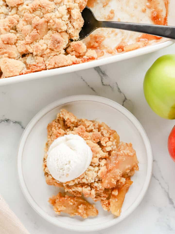 Overhead shot of plated apple cobbler with a scoop of vanilla ice cream on top next to baking dish of cobbler with blacK spoon.