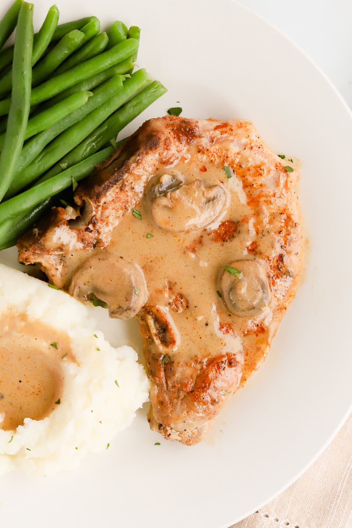 Bone-in pork chop smothered in mushroom gravy on a white plate surrounded by mashed potatoes and green beans.