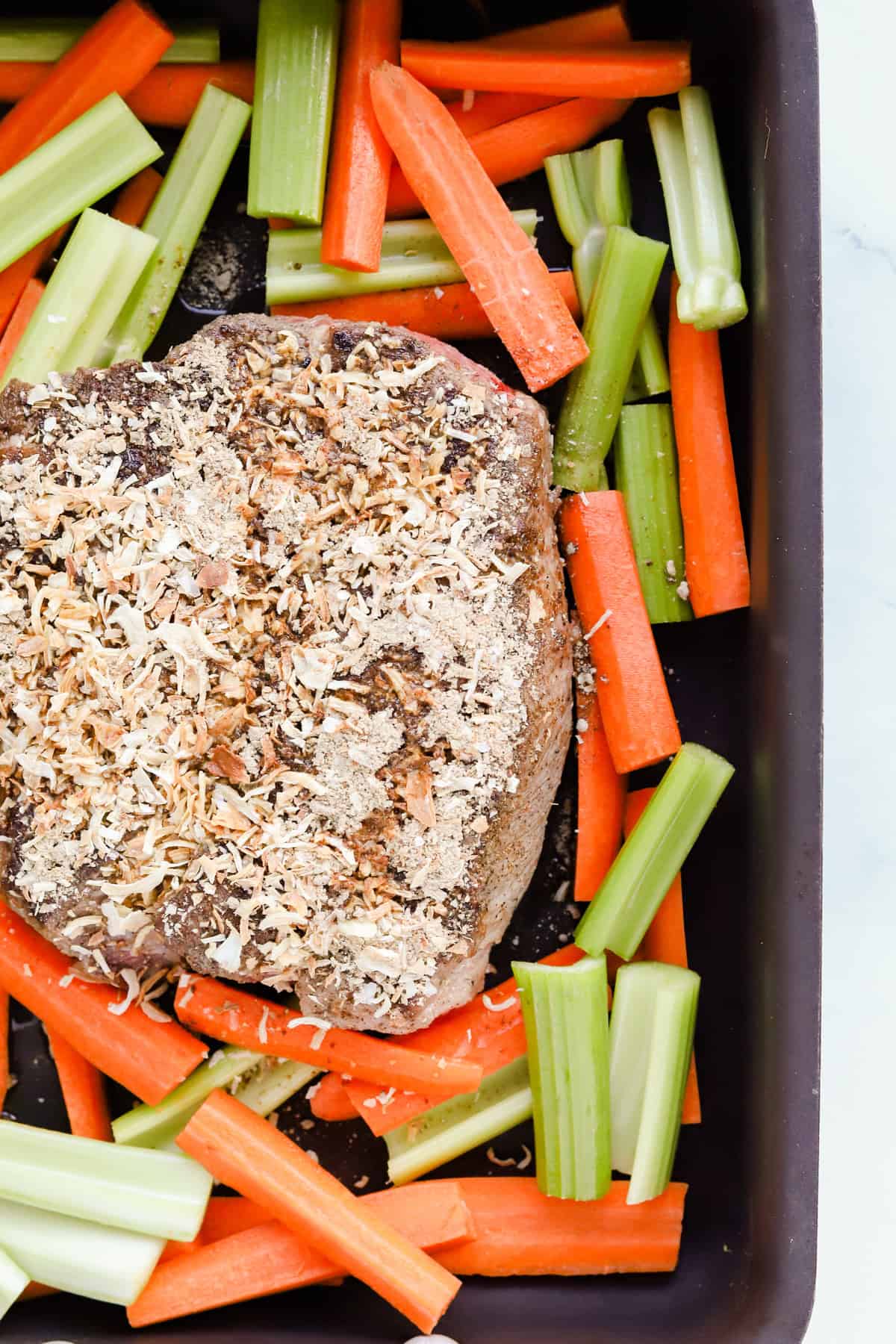 Seared chuck roast topped with vinegar and onion soup mix in a roasting pan surrounded by carrots and celery.