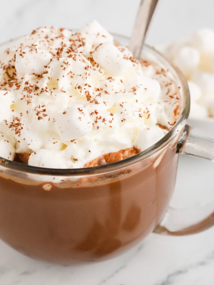 Dark chocolate hot chocolate in clear mug with whipped cream, mini marshmallows and chocolate shavings on top.
