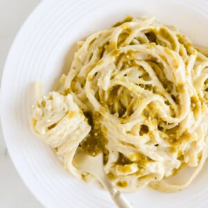 White plate of pesto alfredo pasta garnished with extra pesto and lemon zest.
