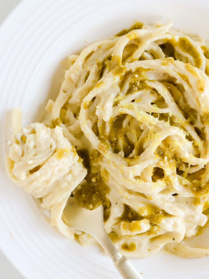 White plate of pesto alfredo pasta garnished with extra pesto and lemon zest.