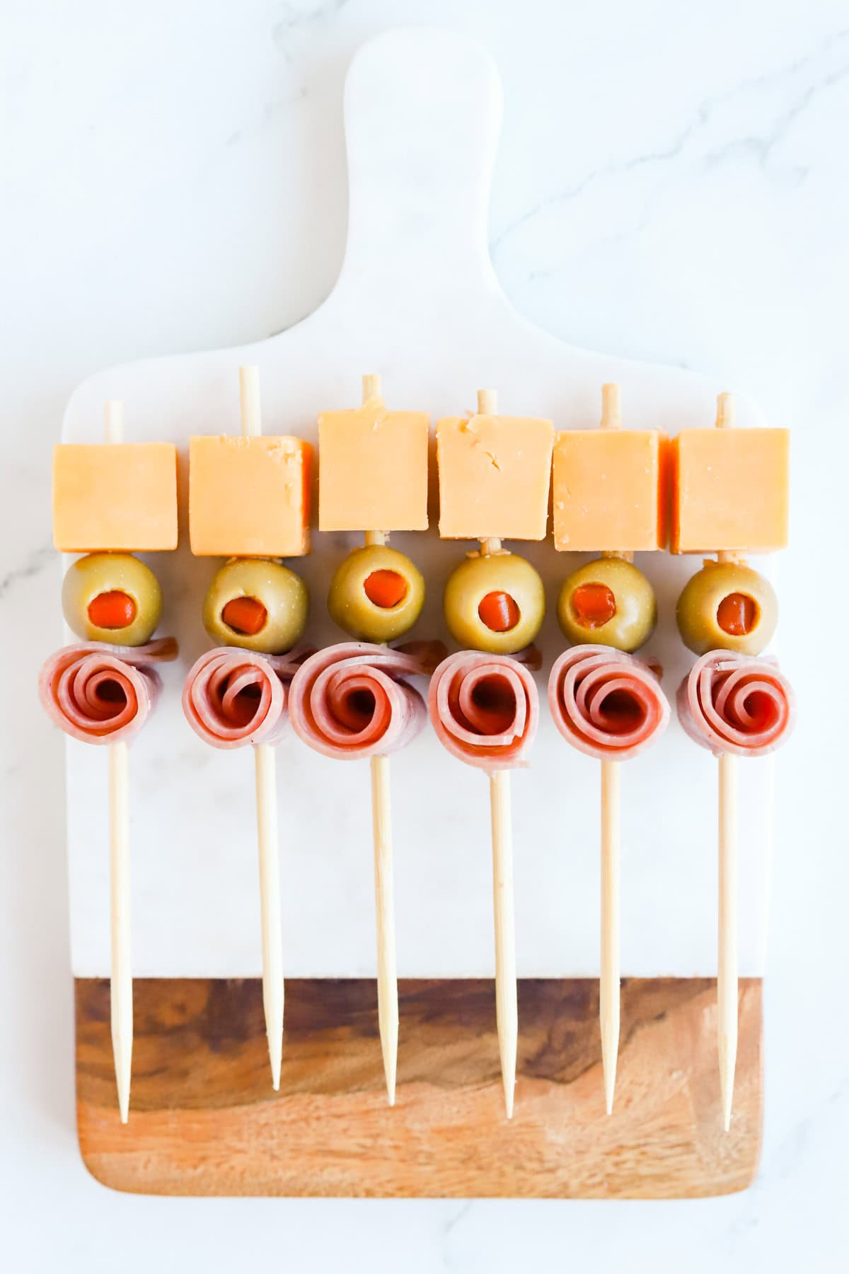 Skewers with cheddar, green olive and salami rose on a marble and wood board.