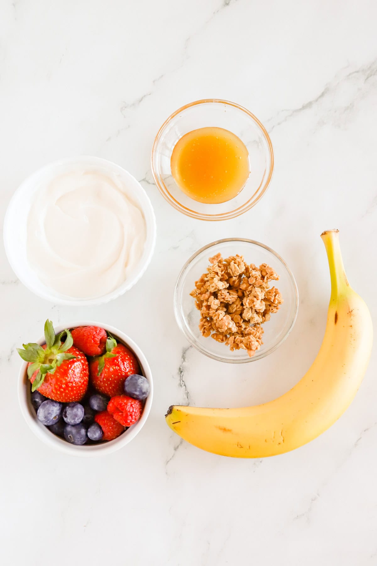 Vanilla yogurt, honey, granola, berries and a banana on countertop.