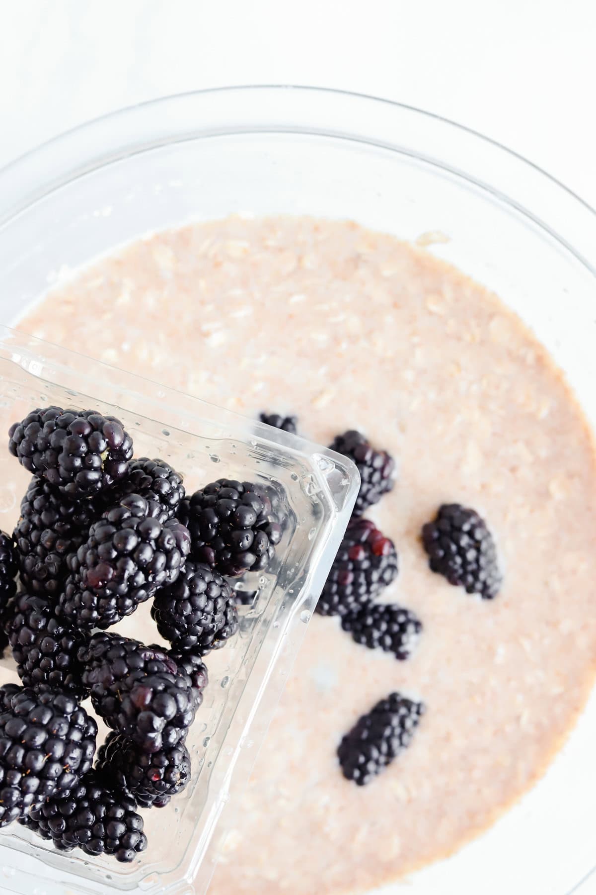 Clamshell of fresh blackberries being dumped into baked oatmeal batter in a glass bowl.