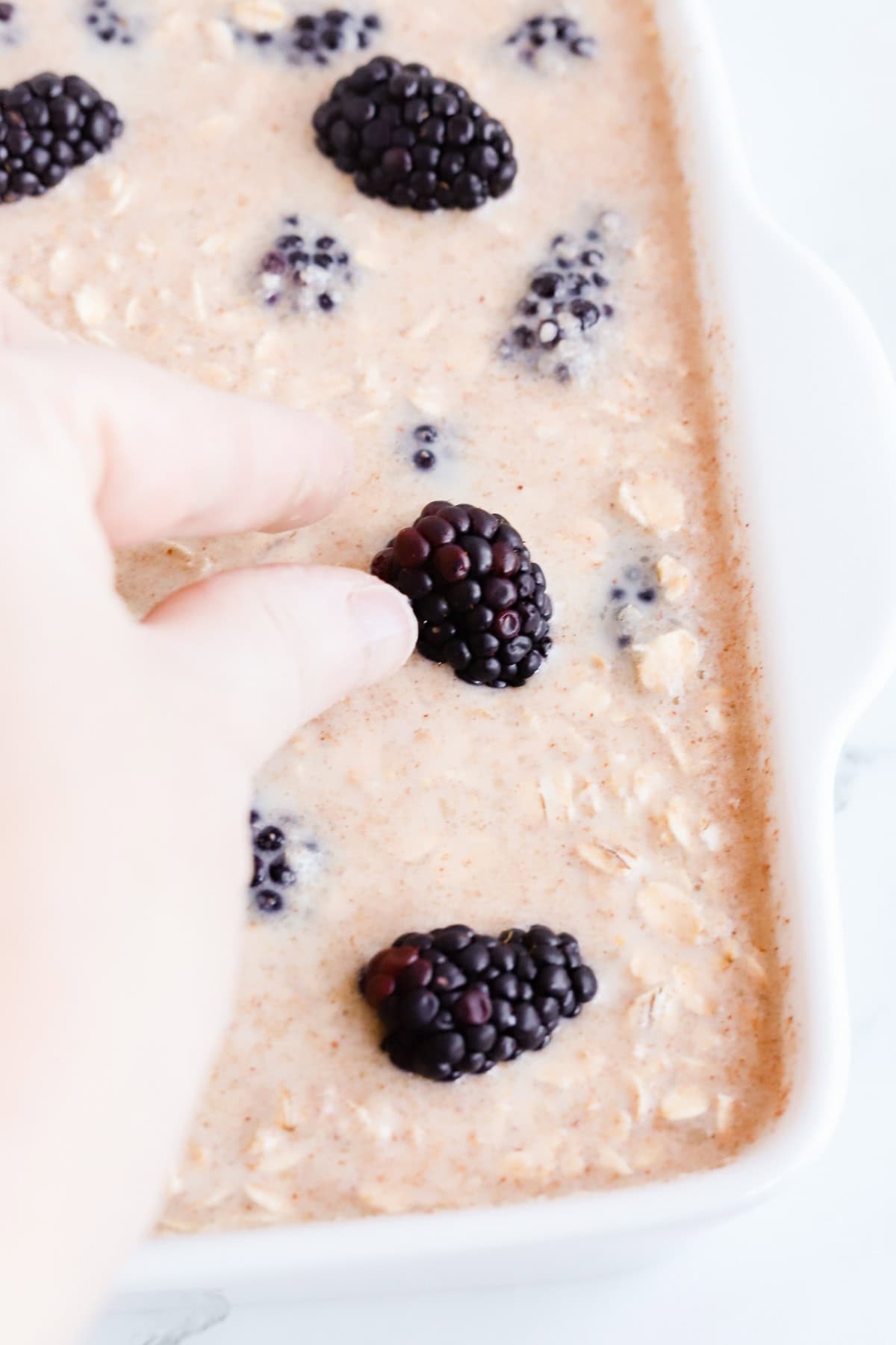 Hand placing fresh blackberries on top of oatmeal in white baking dish before baking.