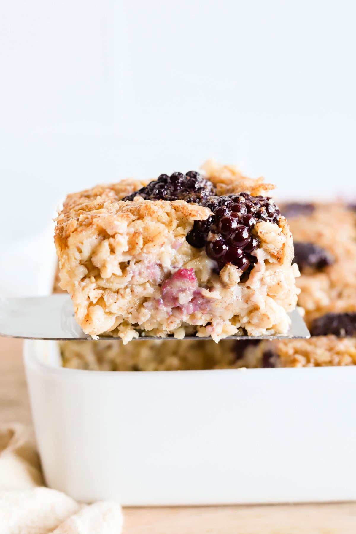 Flat spatula lifting a square of baked oatmeal out of white baking dish.