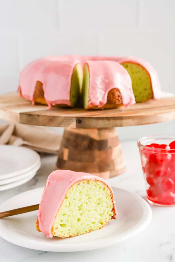 Slice of pistachio cake with cherry glaze on a white plate with whole cake on a wooden cake stand and a glass jar of maraschino cherries.