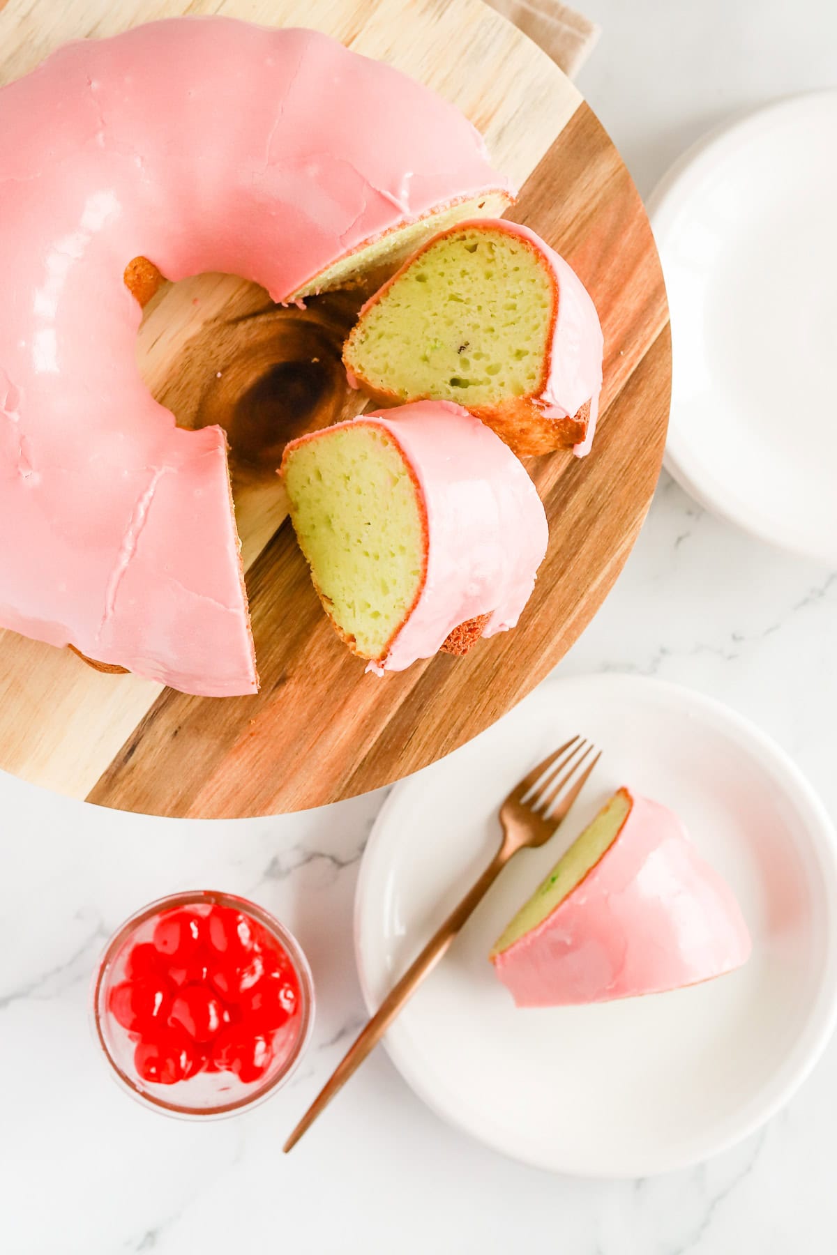 Overhead shot of a slice of pistachio cake on a white plate with a fork next to whole cake on a wooden cake stand.