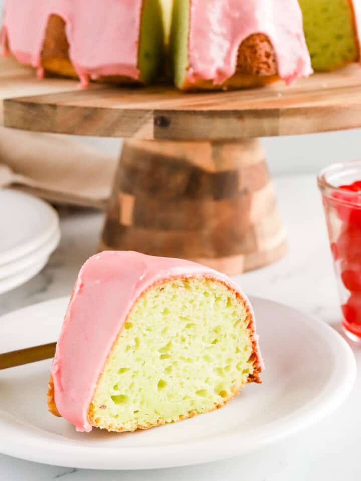 Slice of pistachio cake with cherry glaze on a white plate with whole cake on a wooden cake stand and a glass jar of maraschino cherries.