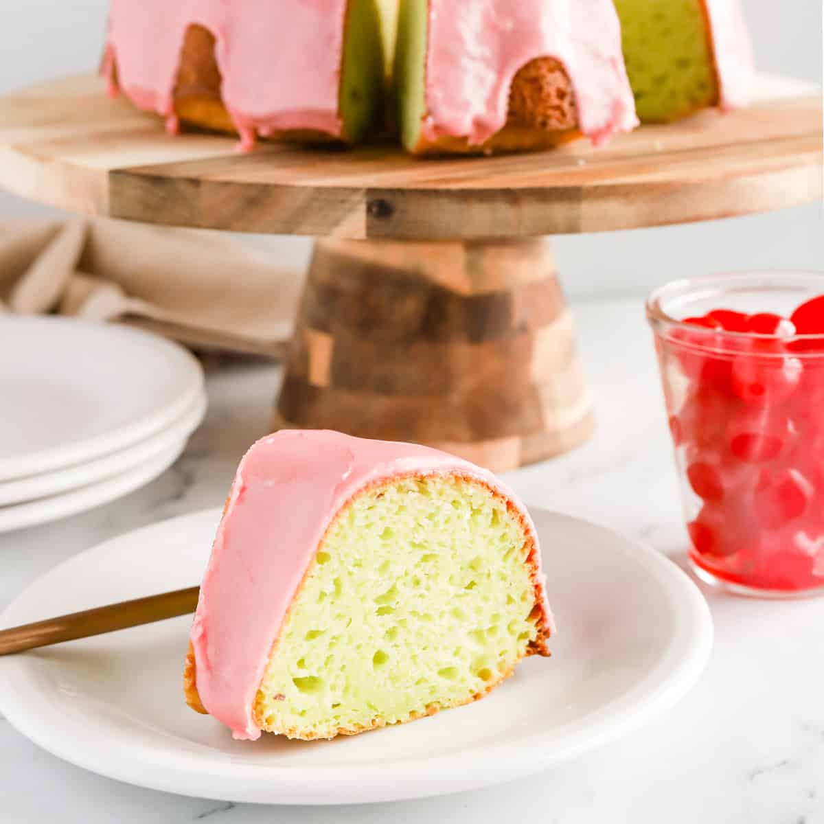 Slice of pistachio cake with cherry glaze on a white plate with whole cake on a wooden cake stand and a glass jar of maraschino cherries.