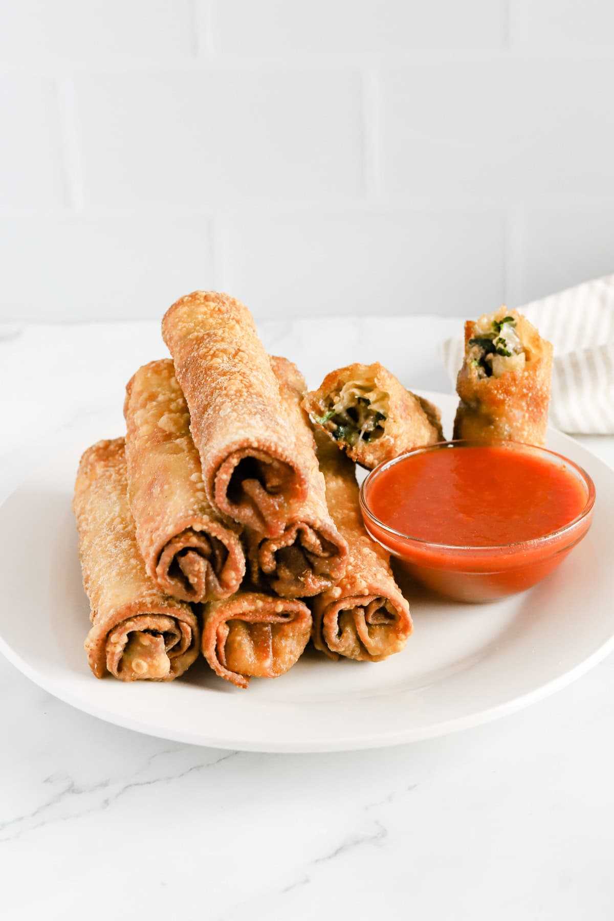 Plate of italian egg rolls stacked next to a glass dish of marinara sauce.