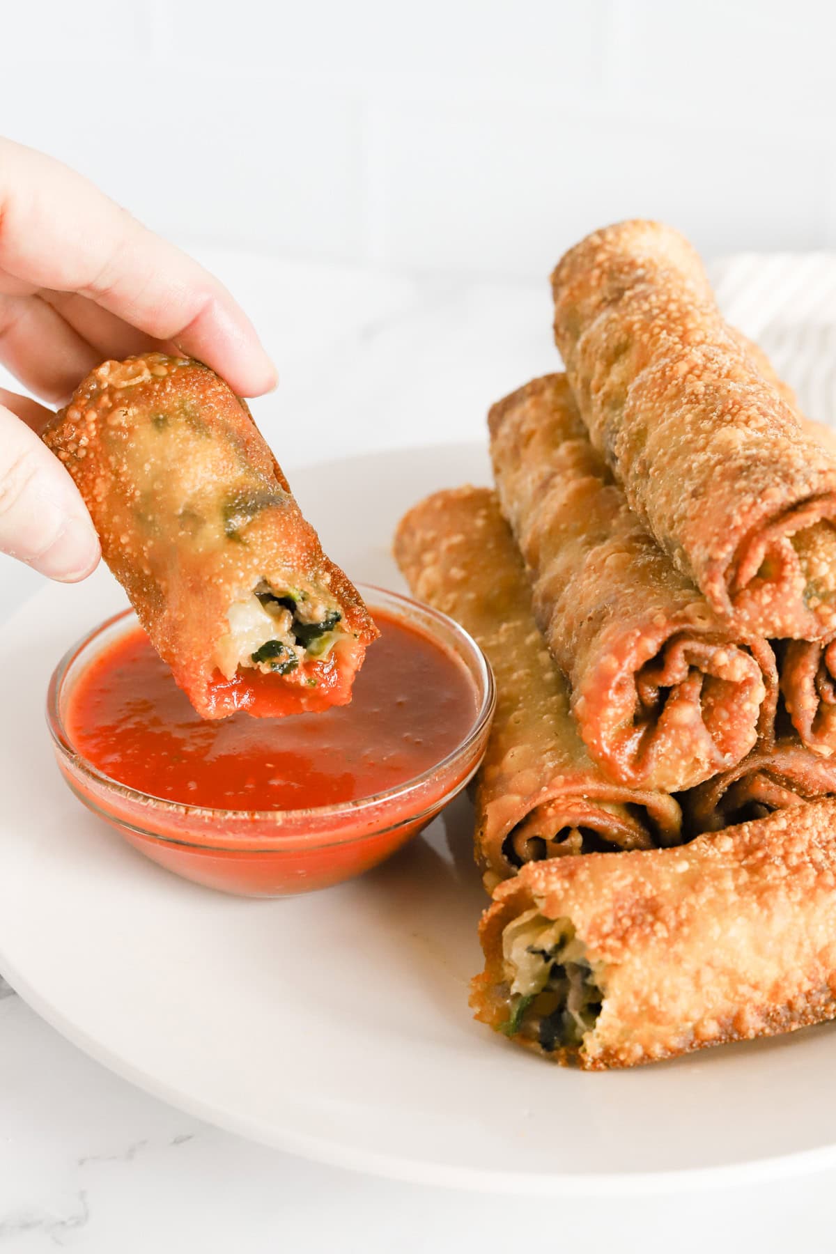 Hand dipping italian egg roll in glass dish of marinara sauce.