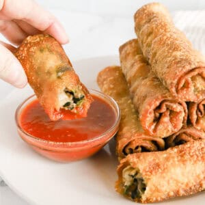 Hand dipping italian egg roll in glass dish of marinara sauce.