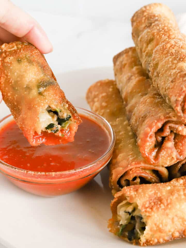 Hand dipping italian egg roll in glass dish of marinara sauce.