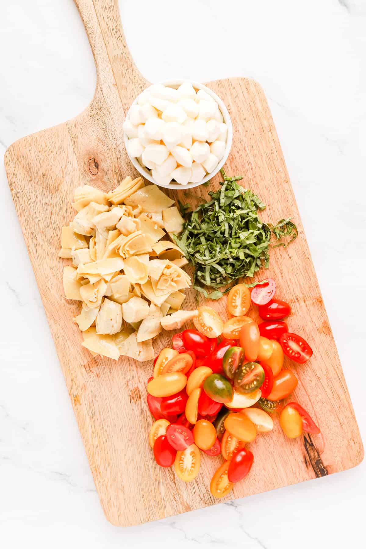 Halved cherry tomatoes, chopped artichokes, chopped basil and mozzarella pearls on a wooden cutting board.