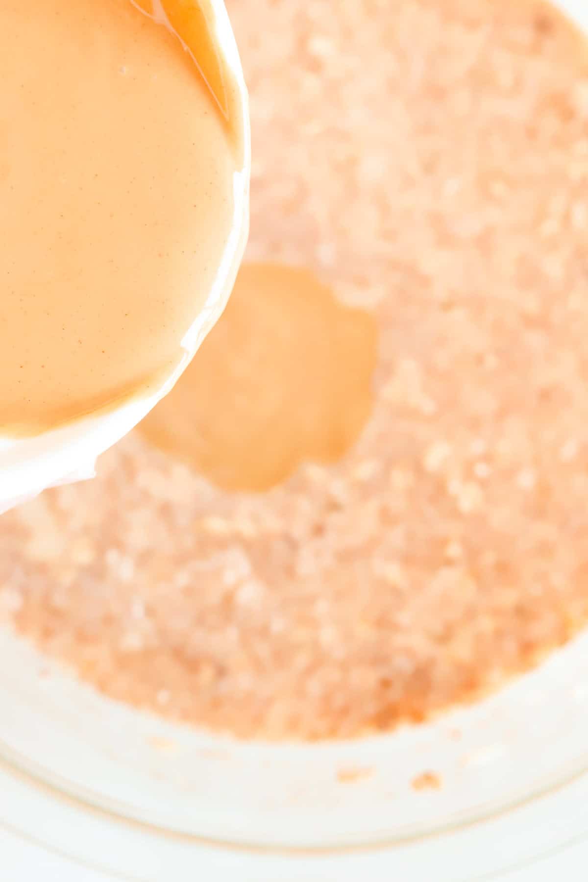 Peanut butter being poured from a white dish into glass bowl of oatmeal batter.