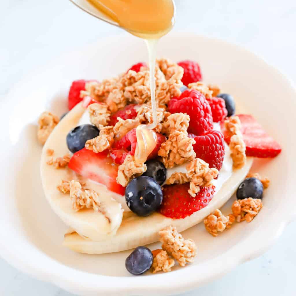 Spoon drizzling honey over breakfast banana split with granola and berries.
