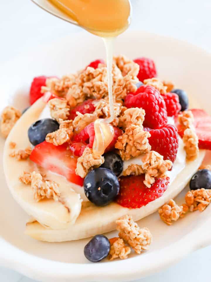 Spoon drizzling honey over breakfast banana split with granola and berries.