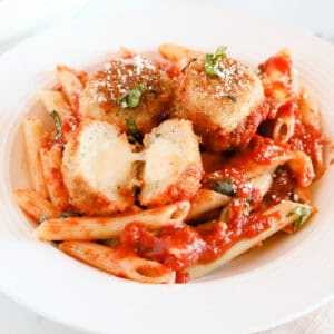 Chicken parmesan meatballs served with penne noodles. One meatball is cut in half exposing the melted mozzarella.