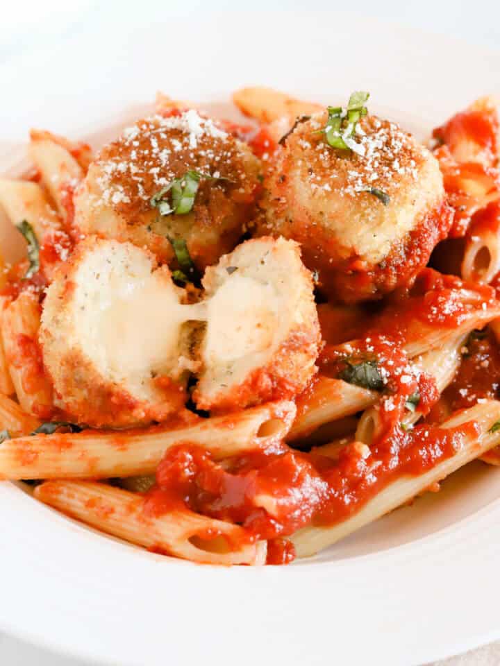 Chicken parmesan meatballs served with penne noodles. One meatball is cut in half exposing the melted mozzarella.
