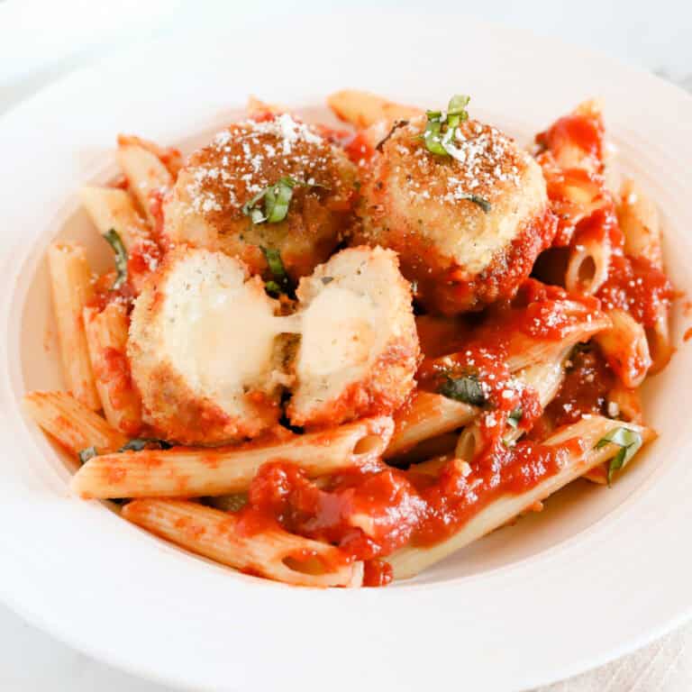 Chicken parmesan meatballs served with penne noodles. One meatball is cut in half exposing the melted mozzarella.